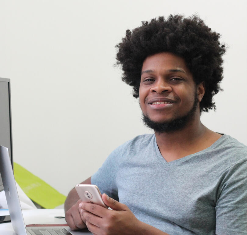 Picture of me at a desk holding a phone with a laptop in front of me. I am looking at the camera smiling. I'm a black man wearing a mid-size afro, small beard and barely no mustache.